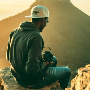 Person sitting down ontop of a rock ledge, with a camera in hand and a mountain vista ahead